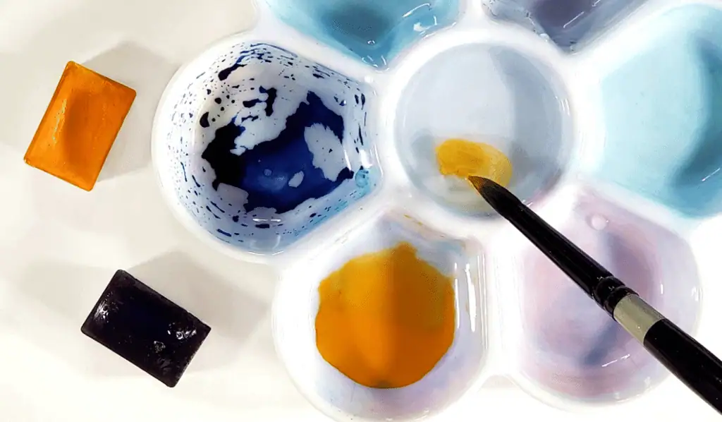 There is a mixing well that's full of pigment as well as a paintbrush that is dipping into the water. In addition, there's one orange watercolor pan and a blue watercolor pan next to it,